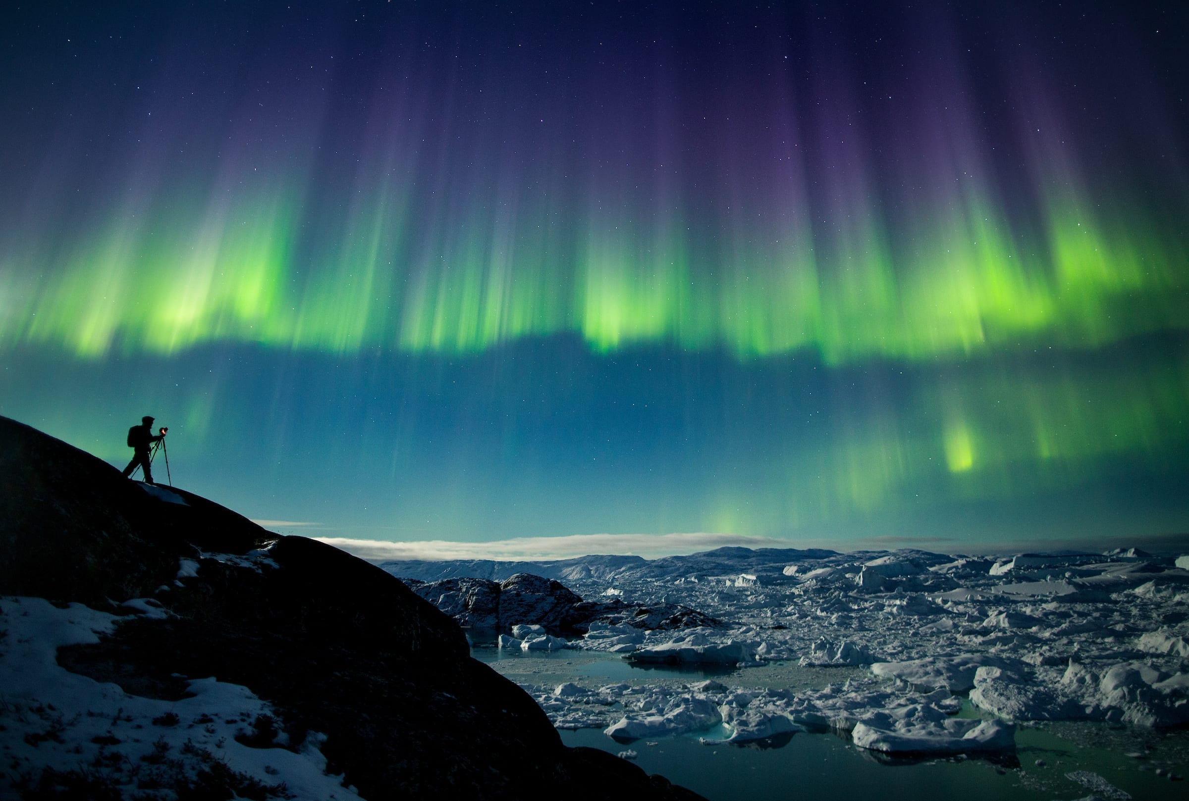 A photographer on a photo tour in North Greenland captures the perfect nighttime shot of northern lights, the starry night sky, and icebergs in the Ilulissat Icefjord